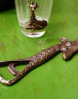 A bronze-colored brass giraffe bottle opener with intricate patterns on a green table next to a shot glass with a giraffe design.