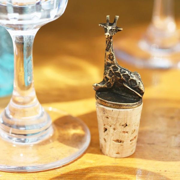A brass giraffe-shaped wine bottle stopper sitting atop corks on a wooden table next to a wine glass.