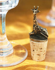 A brass giraffe-shaped wine bottle stopper sitting atop corks on a wooden table next to a wine glass.