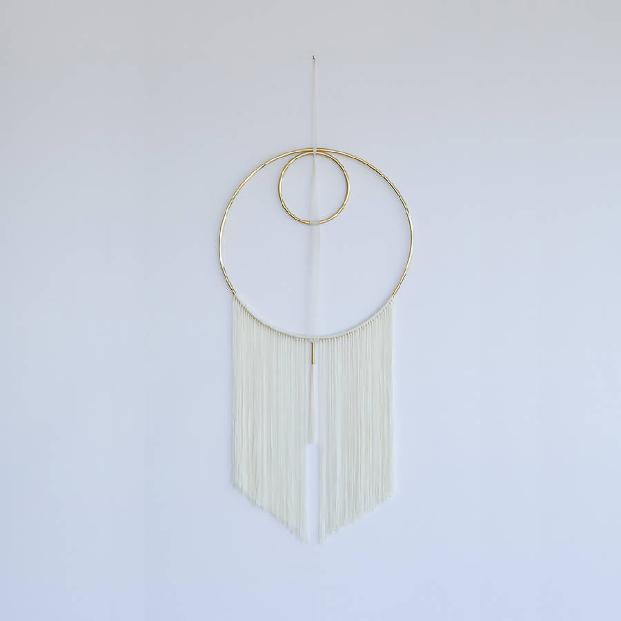A handwoven wall hanging featuring a brass ring and white nylon yarn, with a modern geometric design, displayed against a white background.