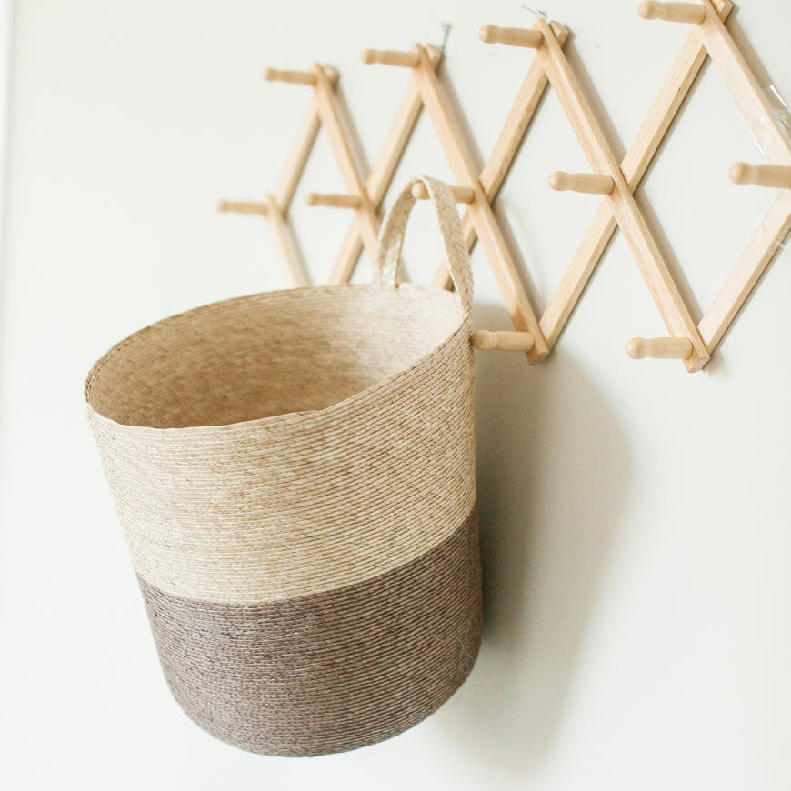 A woven hanging basket in a two-tone color with a decorative expandable storage rack made of bamboo in the background.