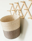 A woven hanging basket in a two-tone color with a decorative expandable storage rack made of bamboo in the background.