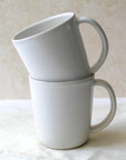Two white ceramic mugs, one stacked inside the other, with a half-moon handle design on a light background