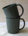 Two green ceramic mugs, one stacked inside the other, with a half-moon handle design on a light background.