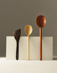 Three handcarved wooden kitchen utensils, including a large spoon, a medium spoon, and a spatula, displayed on a white background
