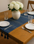 Dining table set with a navy blue woven table runner with decorative tassels, white plates, silverware, and a vase of white flowers.