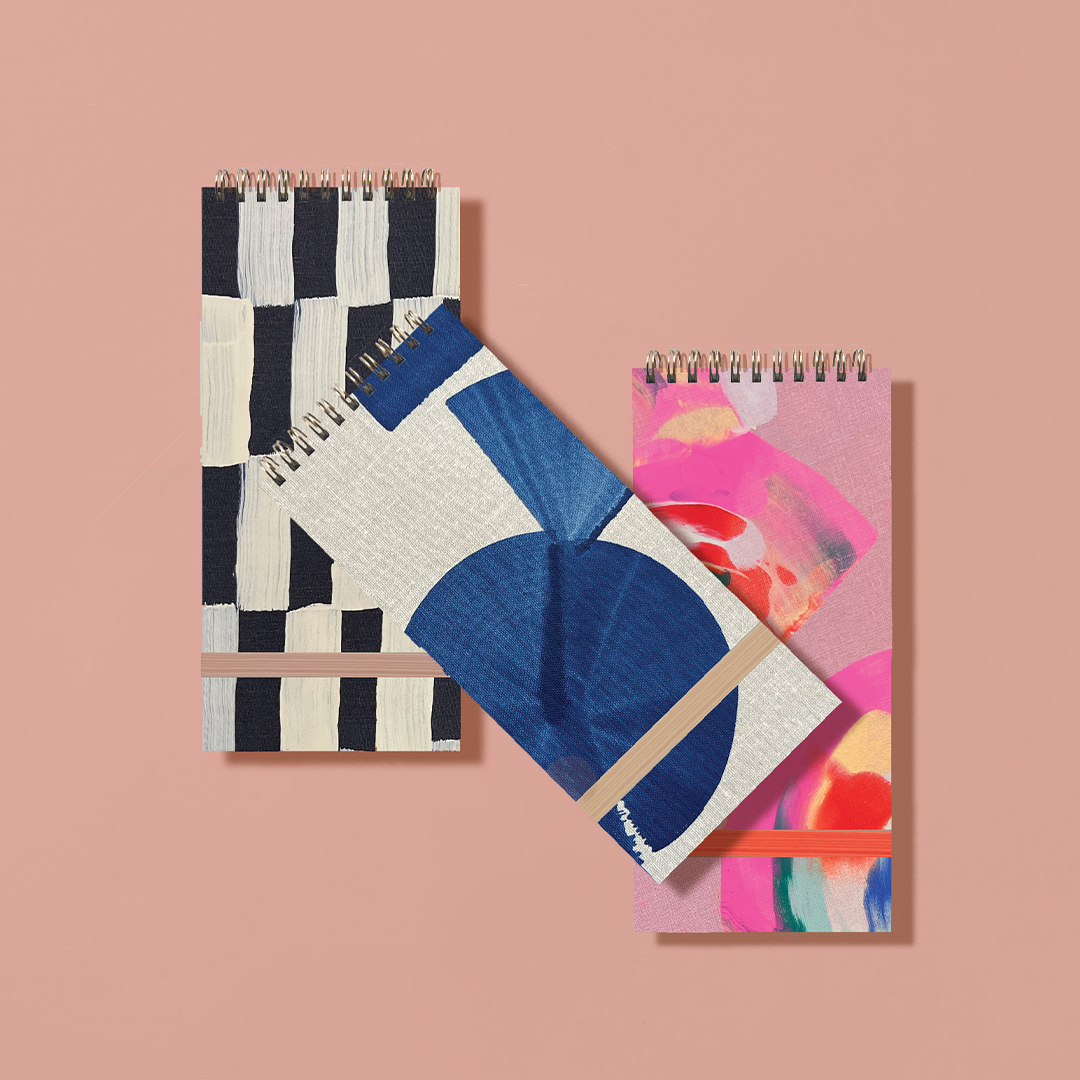 Three spiral-bound notebooks with geometric and abstract designs on a pink background