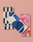 Three spiral-bound notebooks with geometric and abstract designs on a pink background