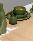 A brown table runner is laid out on a table, with a green vase and various green ceramic plates and cups are placed on top of it