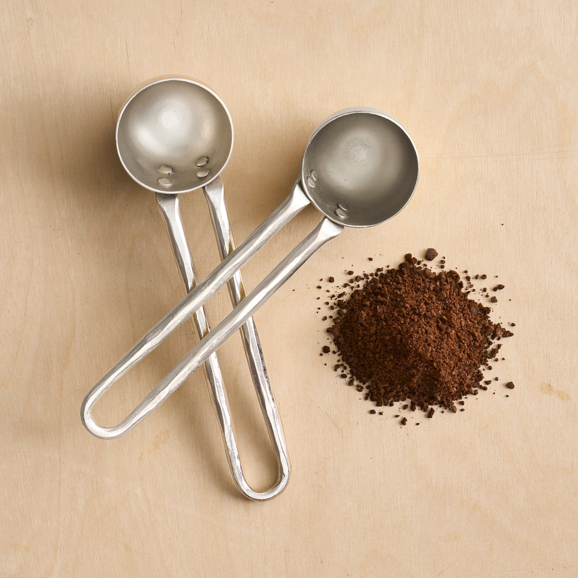Two pewter coffee scoops with a pile of coffee grounds next to them, all placed on a wooden surface