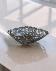 A handcrafted decorative bowl made of assorted recycled iron keys with a copper-colored finish on a gray table with shadows and reflections.