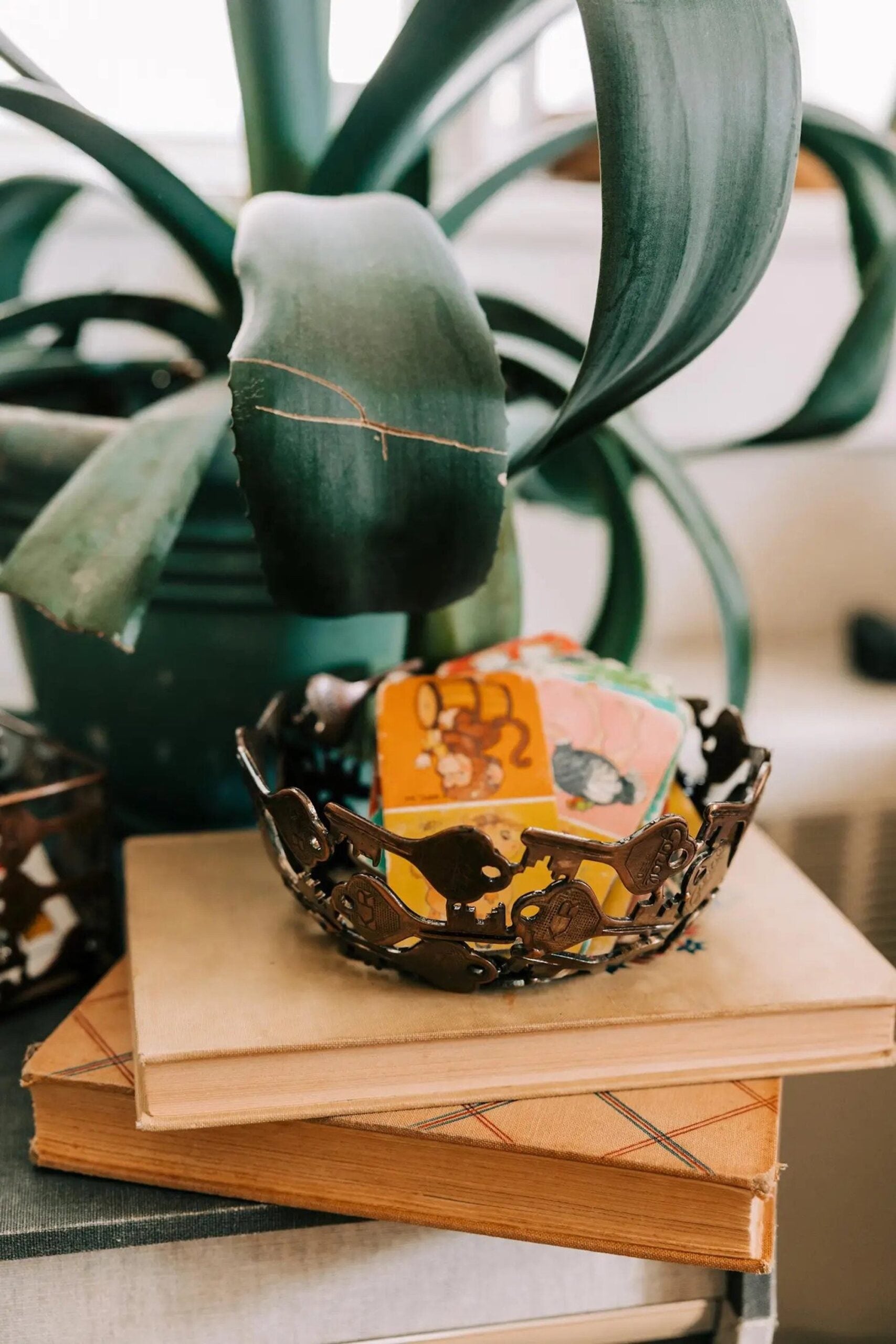 A handcrafted decorative bowl made of assorted recycled iron keys with a copper-colored finish with items inside of it on a wooden table in front of a plant.