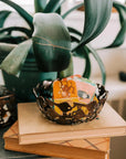 A handcrafted decorative bowl made of assorted recycled iron keys with a copper-colored finish with items inside of it on a wooden table in front of a plant.
