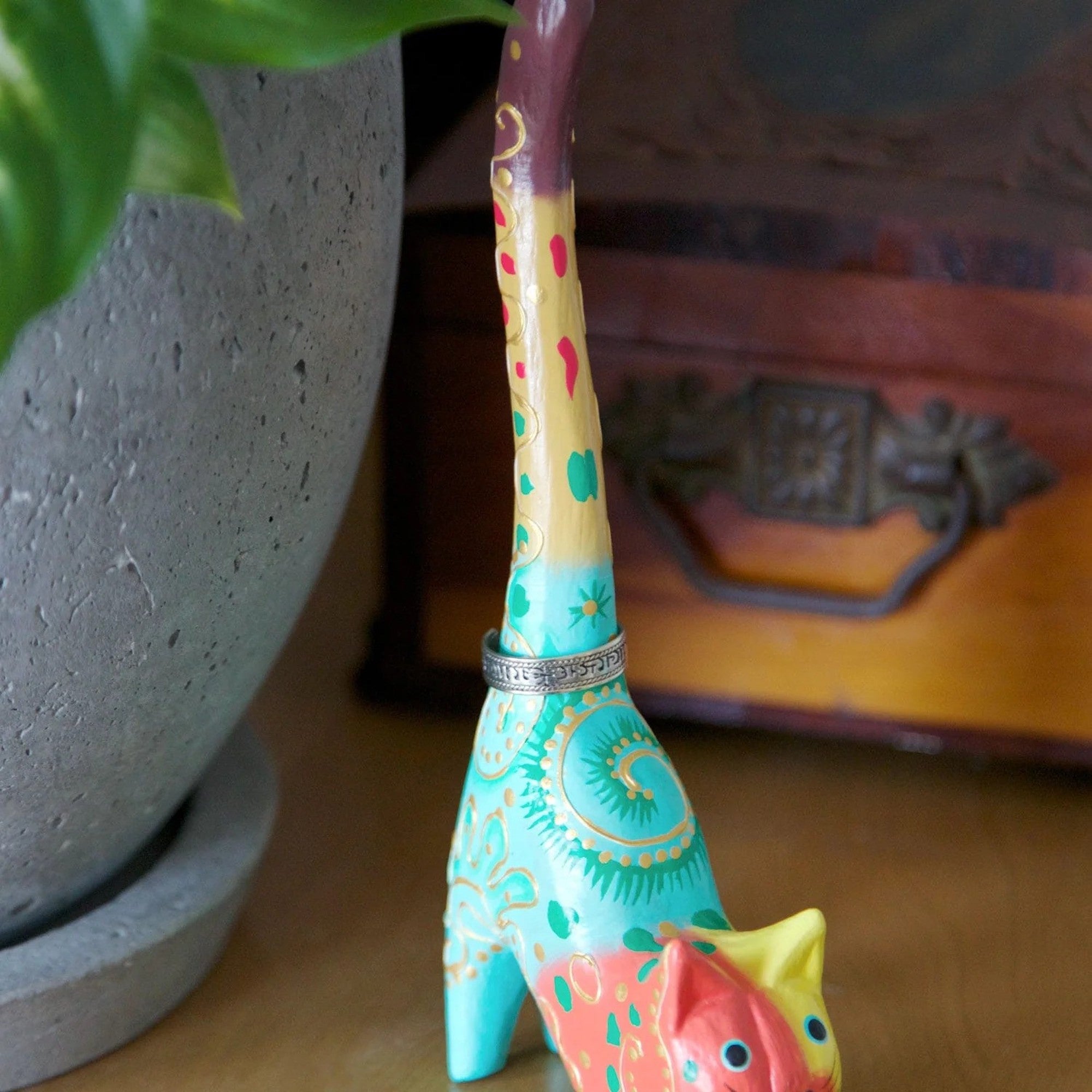 A hand-carved and hand-painted wooden ring holder in the shape of a cat with colorful patterns with a ring on it next to a planter.