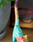 A hand-carved and hand-painted wooden ring holder in the shape of a cat with colorful patterns with a ring on it next to a planter.