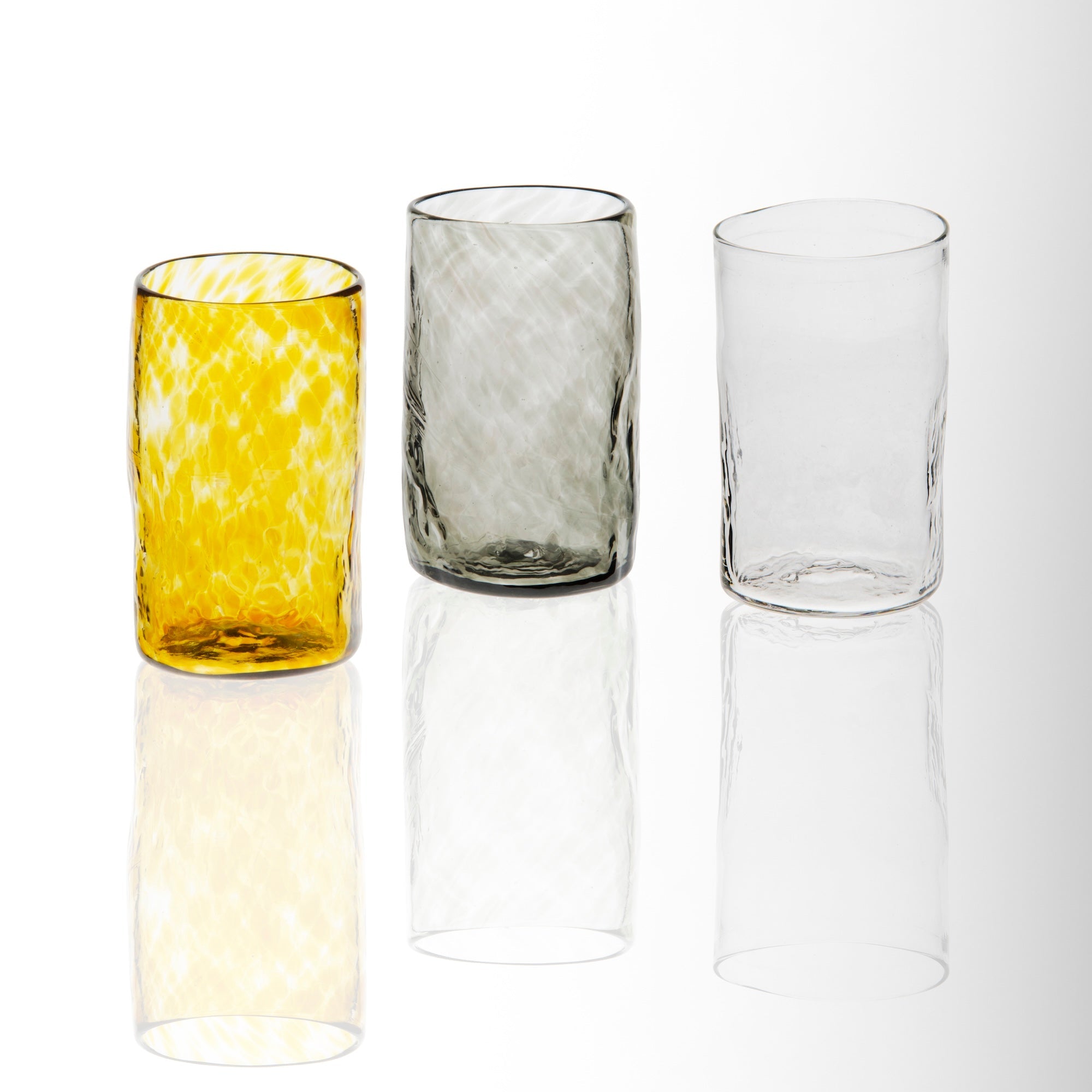 A set of three handblown glass tumblers, with one clear, one amber-colored, and one gray-tinted, displayed against a white background