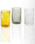 A set of three handblown glass tumblers, with one clear, one amber-colored, and one gray-tinted, displayed against a white background