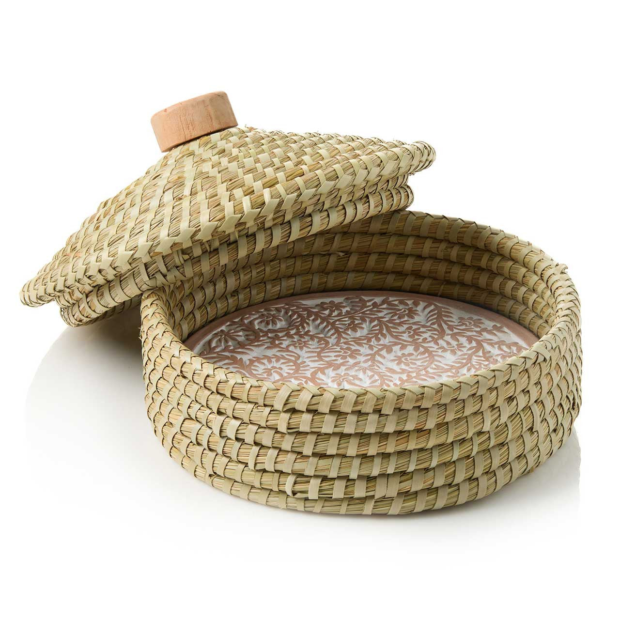 A beige terracotta bread warmer with a pangara design, incised into the stone, sitting within a handwoven kaisa grass basket with a palm wrap detail. A wooden lid is also shown on top.