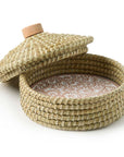 A beige terracotta bread warmer with a pangara design, incised into the stone, sitting within a handwoven kaisa grass basket with a palm wrap detail. A wooden lid is also shown on top.