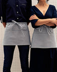 Two people wearing blue waist aprons with multiple pockets and a utility loop