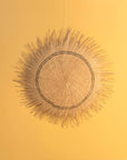 A woven wall decor resembling a sun with a central brown circle and spiky extensions, hanging against a yellow background.