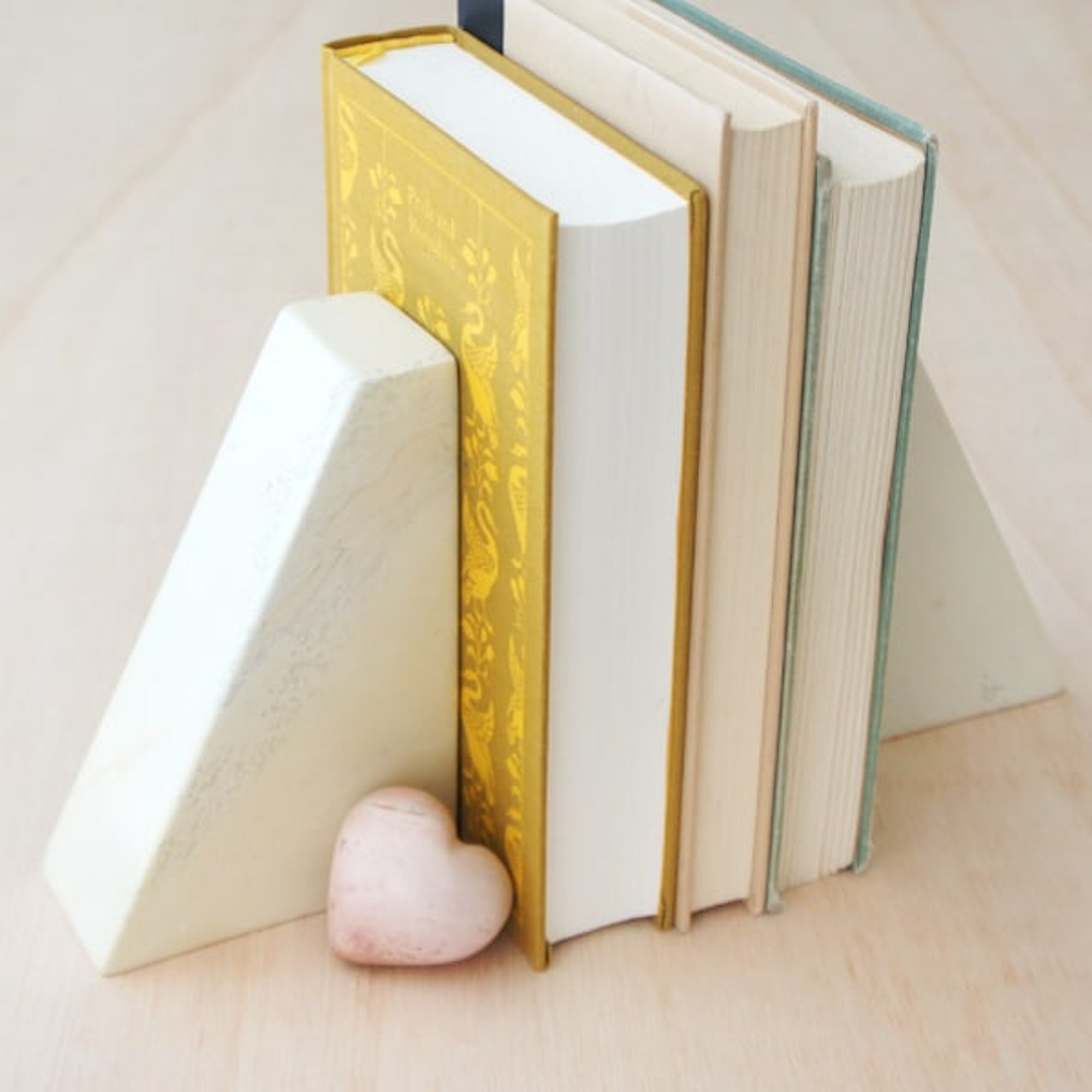 A pair of handmade stone bookends with a pentagonal shape with three books in-between and a heart shaped paperweight next to them