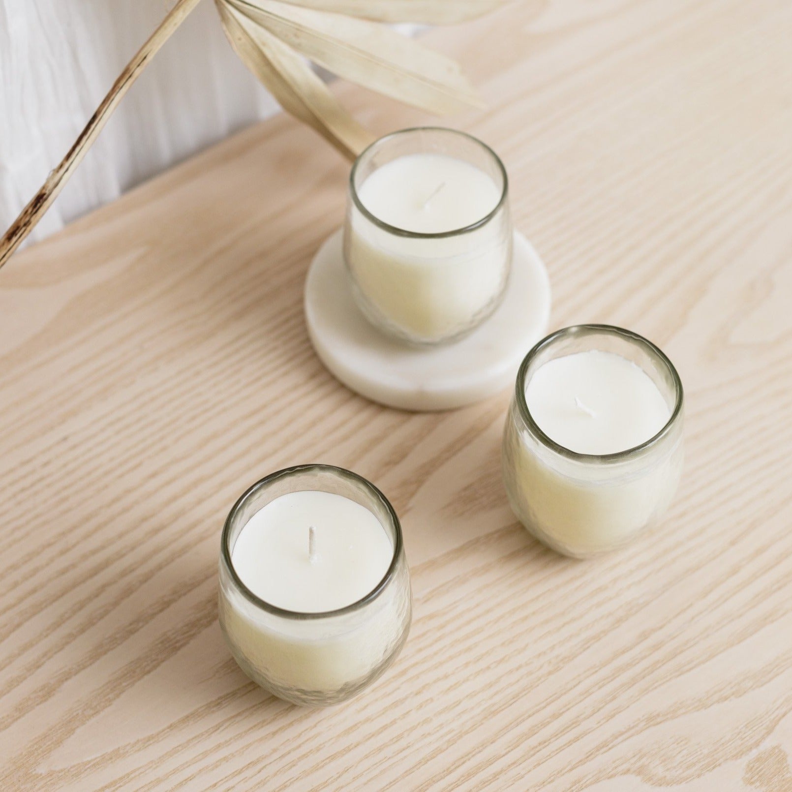 Three white candles in clear glass containers, two on round white holders and one on a flat surface, with a dry plant decoration in the background