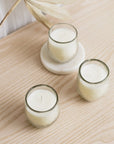 Three white candles in clear glass containers, two on round white holders and one on a flat surface, with a dry plant decoration in the background