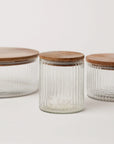 Three fluted glass storage jars in varying sizes with wood lids, the smallest jar is in the front with the largest jar in the background