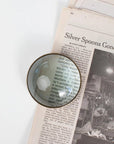 A round paperweight with a magnifying glass in antique brass color, featuring a glass center and a metallic brass outer ring zoomed in on words on top of a newspaper.
