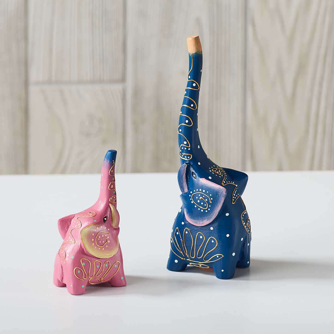 Two decorative elephant figurines, one pink and one blue, on a light surface with a neutral background.
