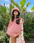 Woman in floral dress and pink hat holding a pink woven bag in a garden setting.