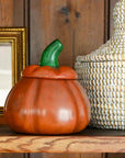 An orange pumpkin-shaped ceramic container with a green stem sits on a wooden shelf next to a woven basket and a gold picture frame against a dark wood paneled wall.