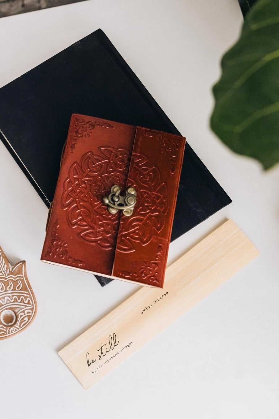 Brown leather embossed journal with decorative clasp on a white surface with a black box and plant leaf.