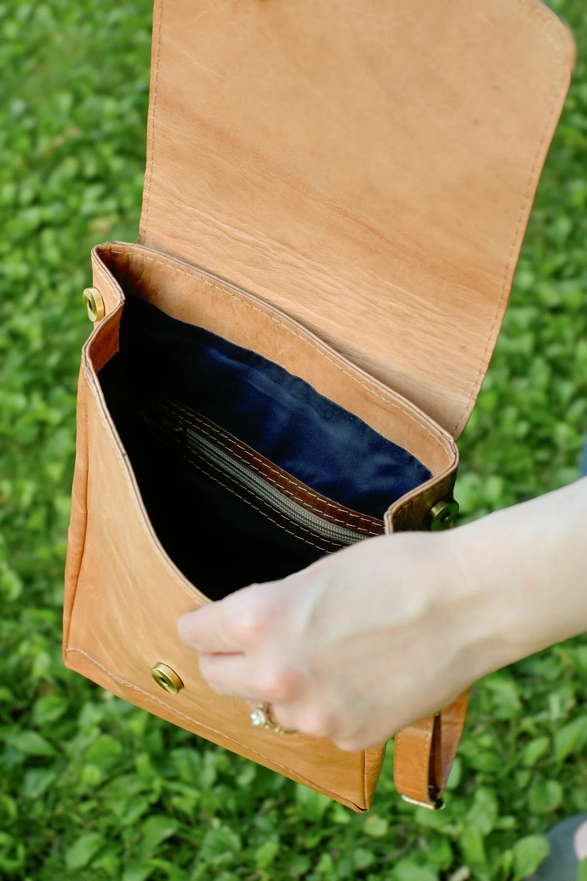 Hand holding a tan leather bag against a grassy background