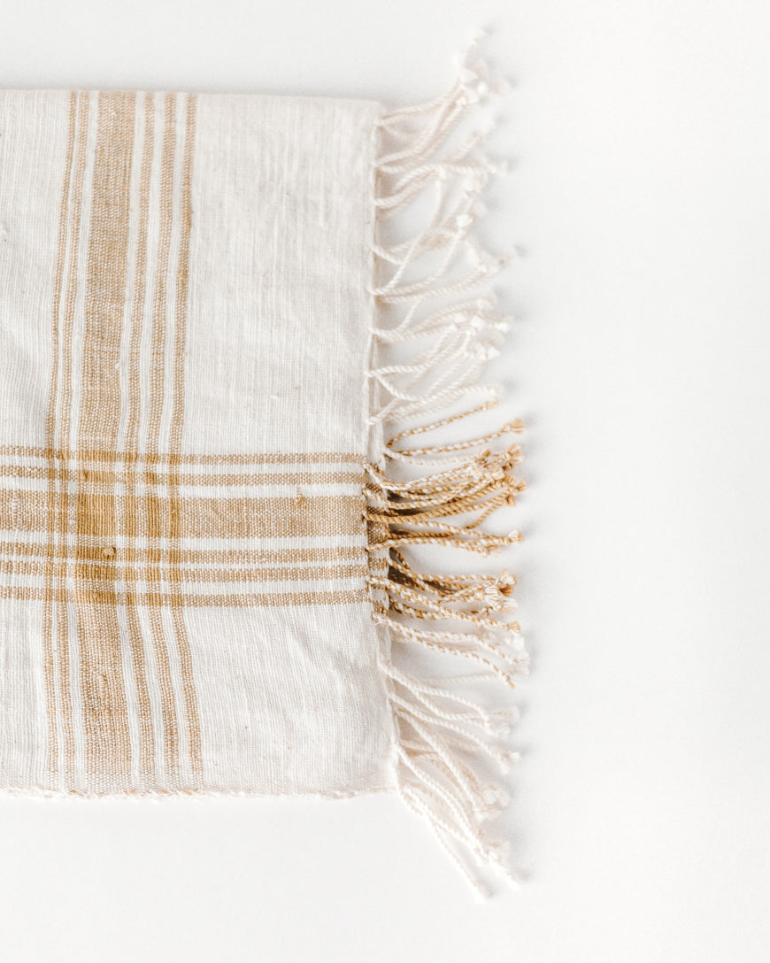 A beige and white striped cotton throw blanket with fringed edges on a light background