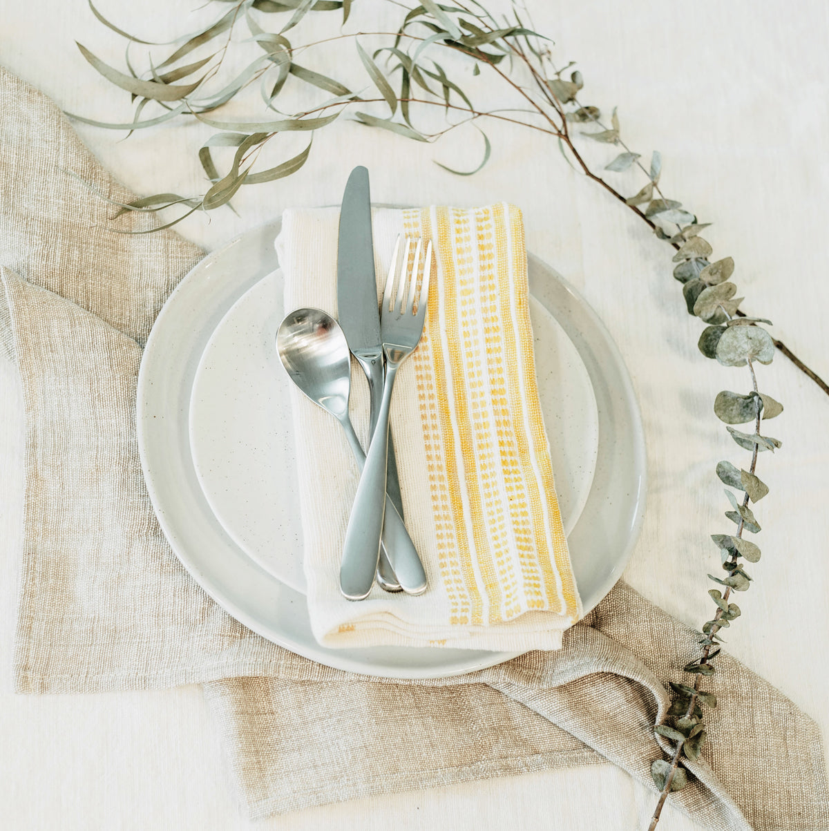 Table setting with plates, cutlery, and a yellow napkin on a textured surface with greenery