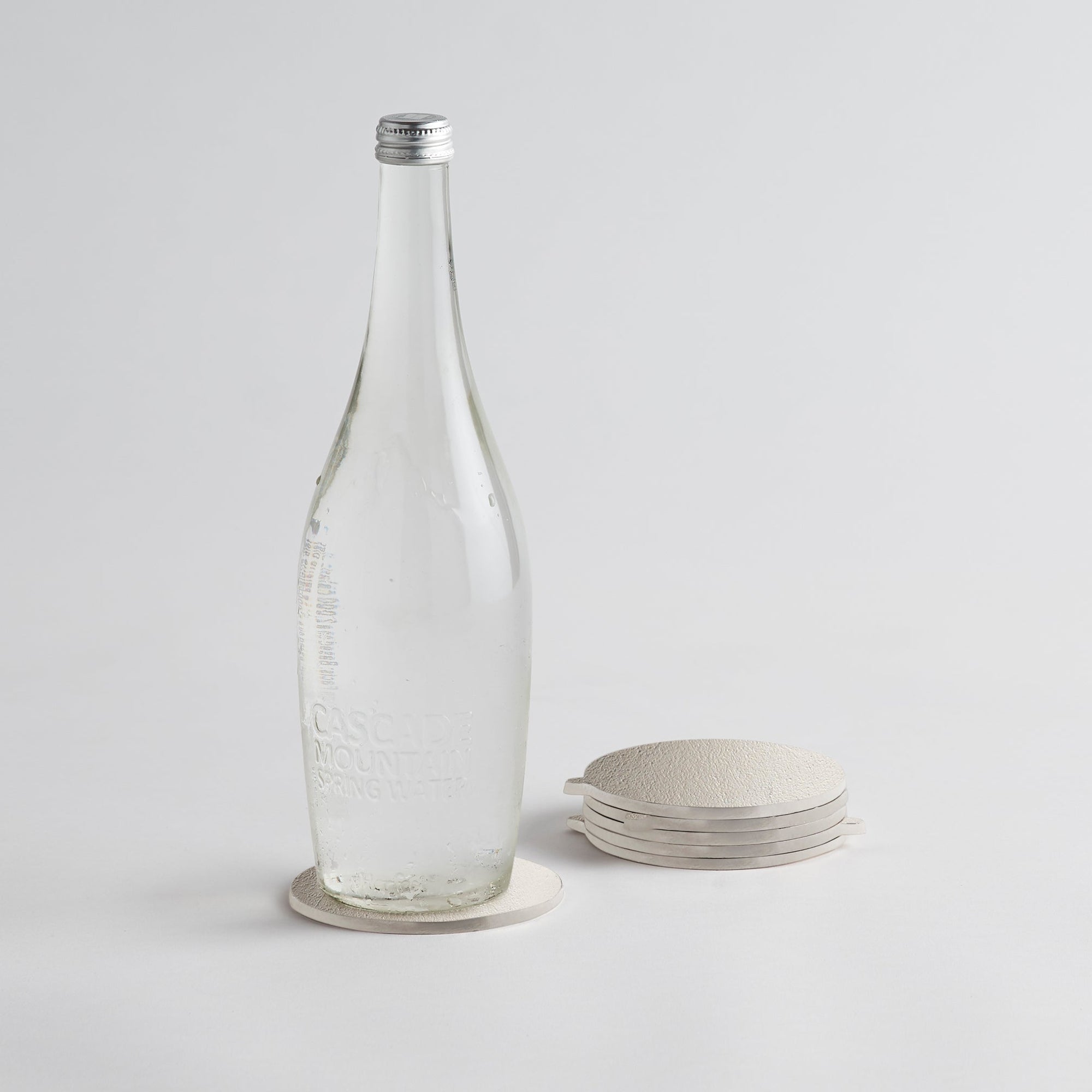 A set of silver cast aluminum coasters with a textured surface, displayed alongside a clear glass bottle