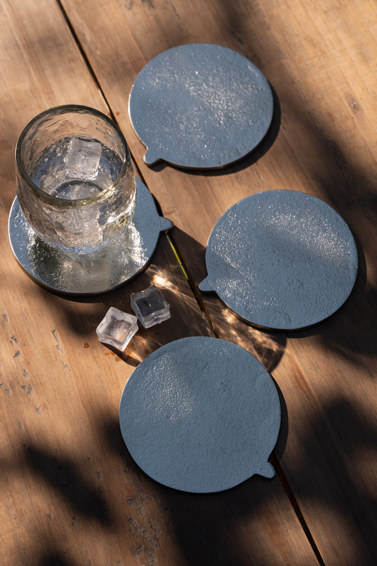 Four silver cast aluminum coasters on a wooden table with a glass of water with ice cubes on top of one of them