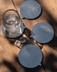 Four silver cast aluminum coasters on a wooden table with a glass of water with ice cubes on top of one of them