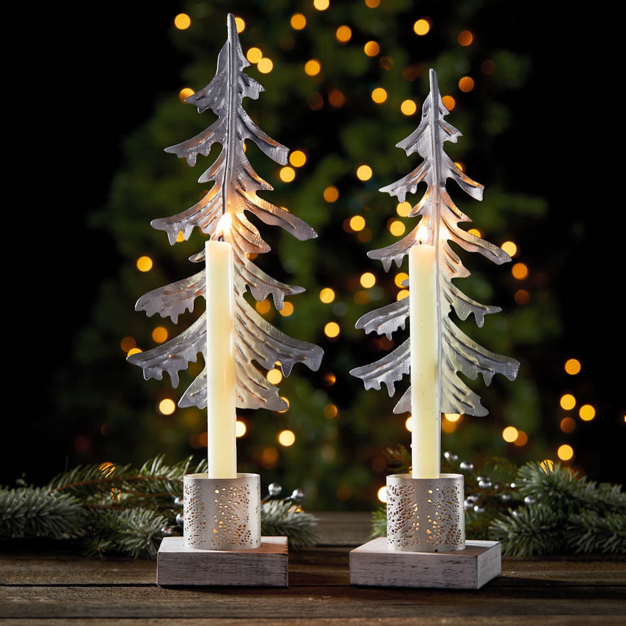 Partially-recycled metal pine in the shape of a tree on whitewashed wooden bases with a small die-cut lantern with lit candles in front of a christmas tree