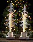 Partially-recycled metal pine in the shape of a tree on whitewashed wooden bases with a small die-cut lantern with lit candles in front of a christmas tree
