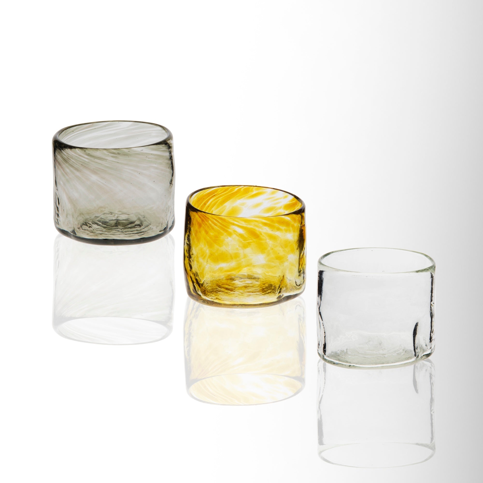 A set of three handblown glass tumblers, one clear, one with a yellow tint, and one with a green swirl, displayed against a white background