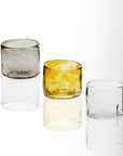 A set of three handblown glass tumblers, one clear, one with a yellow tint, and one with a green swirl, displayed against a white background