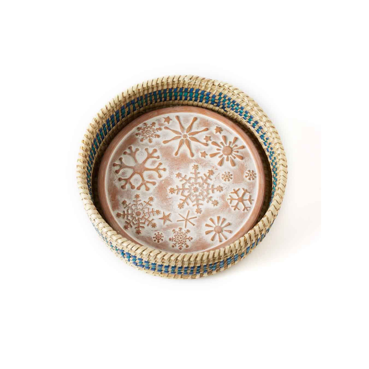 A terracotta bread warmer with a snowflake design, encased in a woven grass basket with blue dyed palm wrap details.
