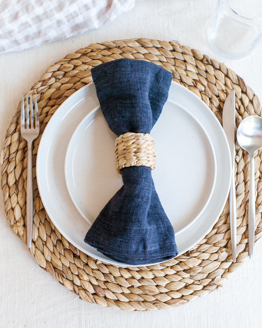 A beige napkin ring holding a blue napkin on top of a white plate and water hyacinth charger