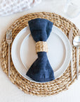 A beige napkin ring holding a blue napkin on top of a white plate and water hyacinth charger