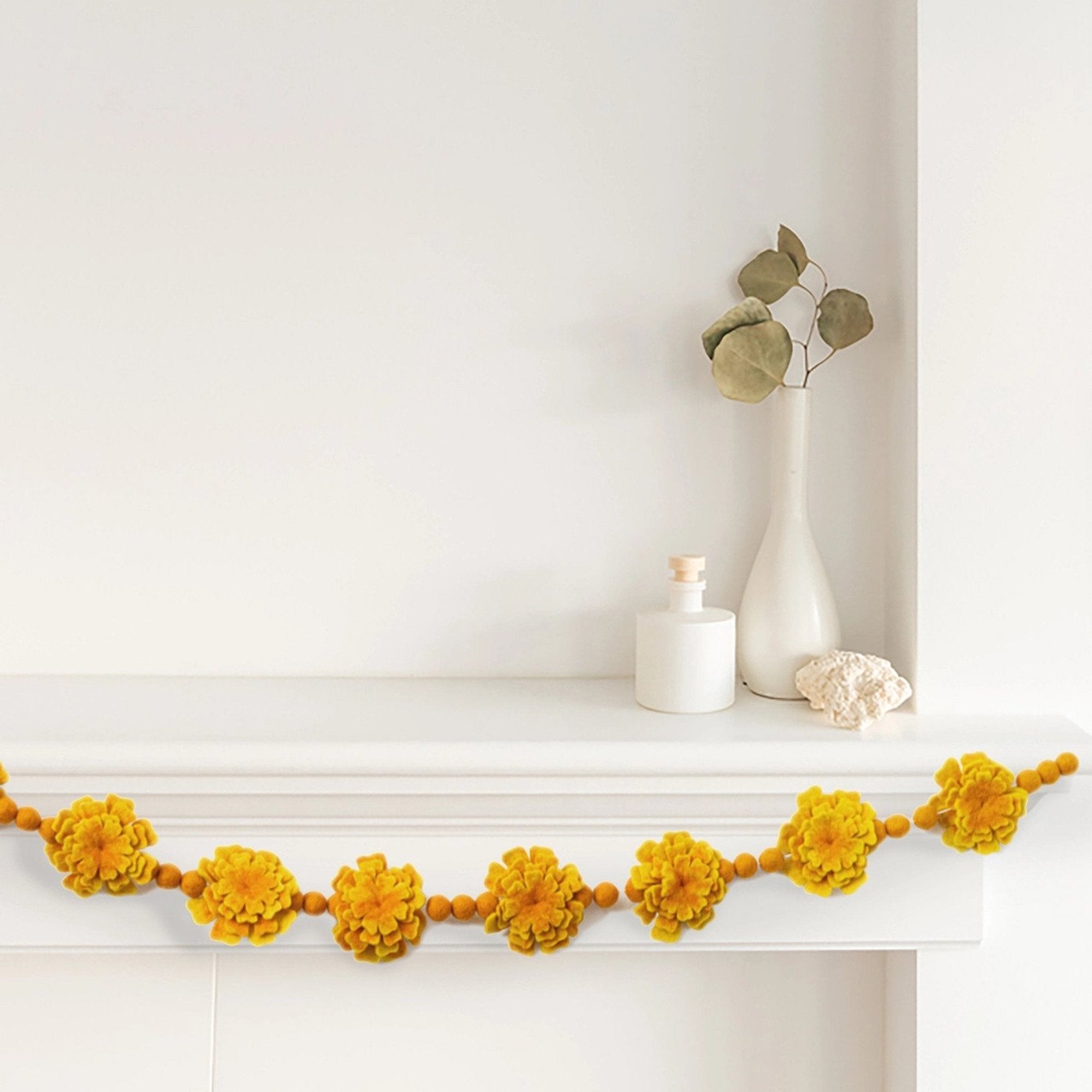 A hanging garland made of felt marigold flowers and beads in yellow and orange colors in front of a mantel