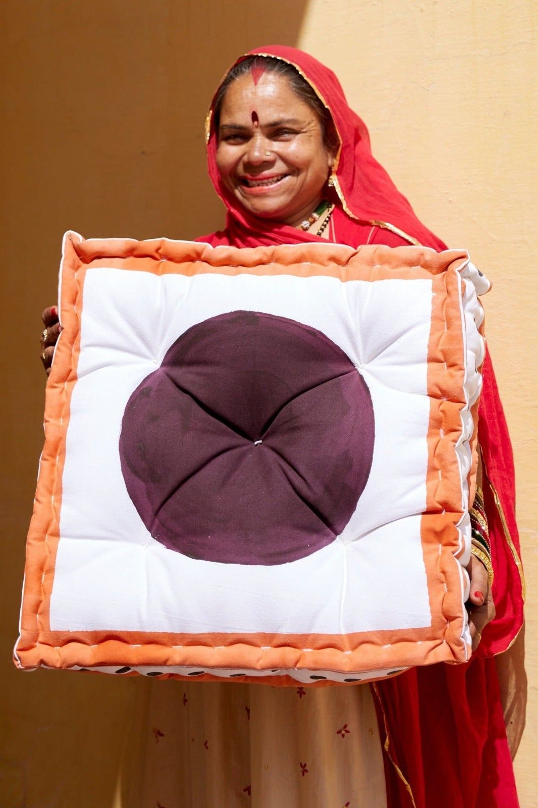 An artisan holding the large cushion with a purple center and orange border that she handmade against a beige wall