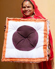 An artisan holding the large cushion with a purple center and orange border that she handmade against a beige wall